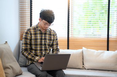 Genç ve çekici Asyalı bir adam evinde rahat rahat dizüstü bilgisayarla çalışıyor. 