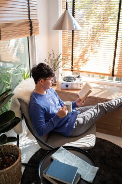 Evinin oturma odasında dinlenirken günlük kıyafetler ve gözlük takan neşeli Asyalı genç adam kitap okuyor.