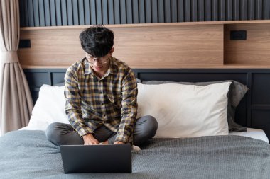 Evden çalışmak, gözlük takan kareli gömlekli bir adam bilgisayar kullanırken yatağa oturur, tüm vücudunu.. 