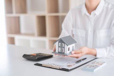 İş için. Beyaz gömlekli adam bir ev modeli taşıyor. Bir masada işlem kağıtları, hesap makinesi, kalem, ev anahtarı ve banknotlar vardır.. 