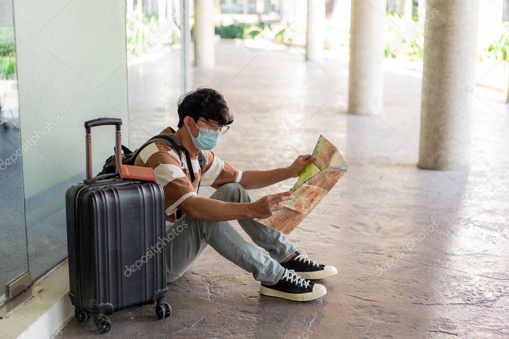 Travel, A man wearing glasses with a striped shirt and mask sit on the floor, reading map in his hand with suitcase placed on right side and backpack.