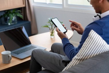 Yakışıklı Asyalı gözlüklü genç adam oturma odasındaki cep telefonuyla interneti tarıyor. 