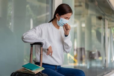 Seyahat et. Uzun kollu beyaz gömlek ve kotlu hasta bir kadın, maske takıyor ve öksürüyor, sağ tarafında bavul, harita, not defteri ve sırt çantasıyla sandalyeye oturuyor..
