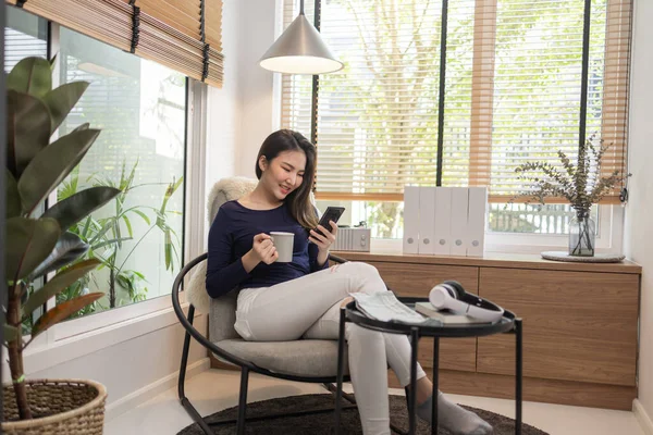 Work from home concept a confident woman sitting on a modern chair hold a cup of coffee and another hand typing a keyboard on her smart phone.