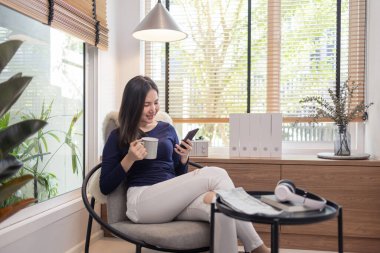 Ev kavramından yola çıkarak modern bir sandalyede oturan kendine güvenen bir kadın bir fincan kahve ve akıllı telefonunda klavye yazan başka bir kadın..