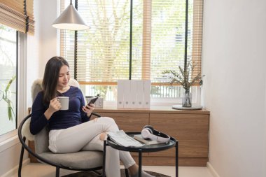 Ev kavramından yola çıkarak modern bir sandalyede oturan kendine güvenen bir kadın bir fincan kahve ve akıllı telefonunda klavye yazan başka bir kadın..