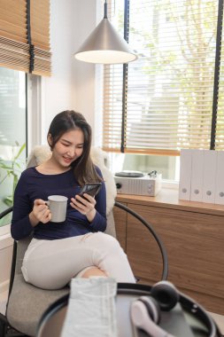 Ev kavramından yola çıkarak modern bir sandalyede oturan kendine güvenen bir kadın bir fincan kahve ve akıllı telefonunda klavye yazan başka bir kadın..