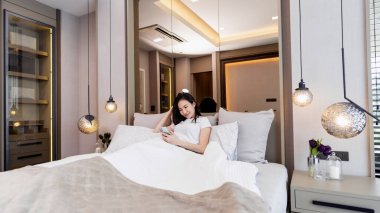 Yatak odası konsepti. Uykulu hisleri olan bir kız. Sırtını birkaç büyük ve rahat yastığa yaslayıp haber bülteni için mobil ekrana bakıyor..