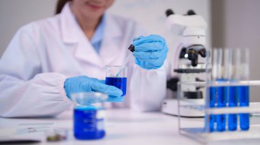 Yetişkin Asyalı bilim kadını modern bir bilim laboratuvarında çalışıyor. Önemli araştırmalar için mavi sıvıyı bir damlatıcı ve beher ile tutuyor..