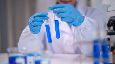 Yetişkin Asyalı bilim adamı test tüplerindeki mavi sıvıyı eldivenli ellerle inceleyerek bilimsel deney ve kimyasal analiz yapıyor..