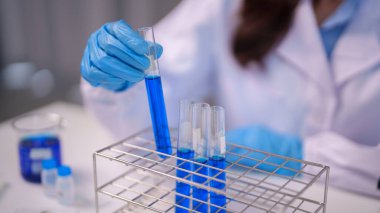 Modern bir laboratuvar ortamında çalışkan kimyasal araştırmalar yapan yetişkin bir bilim adamı. Mavi eldiven, metal raflara yerleştirilmiş canlı mavi bir sıvıyla test tüpünü tutuyor..