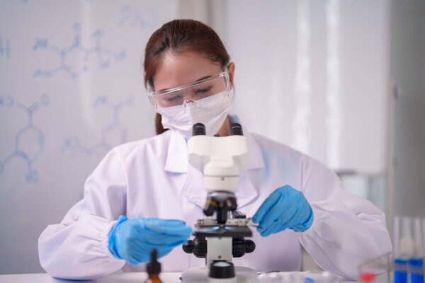 A focused Asian adult female scientist examines specimens through a microscope She works diligently in a modern laboratory environment Chemical structures adorn the background