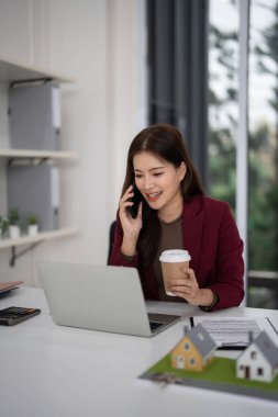 Asyalı emlakçı müşterileri birbirine bağlar. Kadın emlak satışlarını telefonla tartışıyor, dizüstü bilgisayarla çalışıyor. Modern ofis masasında ev modelleri gösteriliyor.