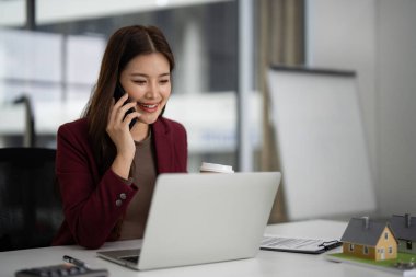 Asyalı profesyonel müşterileri mülkiyet hakkında bilgilendirir ve ev satışları için dizüstü telefonu kullanır. Modern ofis çalışma alanı.
