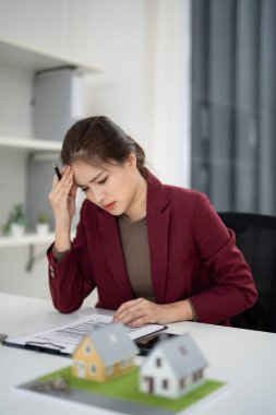 Asyalı kadın ajan, masa başı iş kararlarında ev modelleriyle emlak sözleşmesi belgelerinin incelenmesinde stres hissediyor.