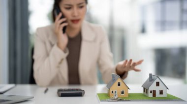 Bulanık geçmişi olan Asyalı yetişkin profesyonel konut piyasası seçeneklerini tartışıyor. Ön planda minyatür evlerin hesap makinesinin anahtarları var. Uzman emlak kararları.