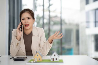 Profesyonel Asyalı ajan cep telefonuyla iletişim kuruyor. Masada ev maketleri ve hesap makinesi var. Karmaşık bir satışla ilgili belirsizlik gösteren bir kadın.