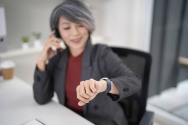 Olgun Asya iş görüşmeleri cep telefonu kontrolleri bilek aygıtı zamanı gösterişli ofis arka planında bulanık iletişim