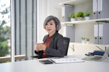 Deneyimli Asyalı yaşlı yönetici parlak ofis masasında kahveyle rahatlıyor. Liderlik ve kurumsal sağlığını gösteriyor..