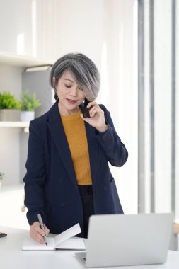 Odaklanmış Asyalı yaşlı yönetici kadın, güneşli bir çağdaş çalışma alanında dizüstü bilgisayarla akıllı telefon yazma stratejisi hakkında konuşuyor