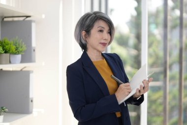 Olgun Asyalı kadın profesyonel, parlak çağdaş çalışma ortamı içindeki bir defterde fikirleri özenle yakalıyor.