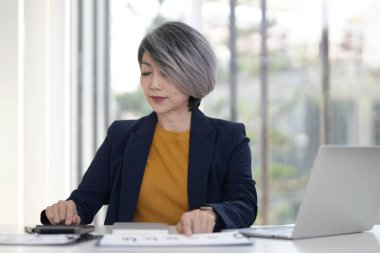 Odaklanmış üst düzey Asyalı kadın, hesap makinesi kullanarak, masa başı iş ortamı veri analizi konseptini kullanarak finansal tablolarda çalışıyor.