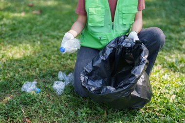 Çevre temizliğini ve geri dönüşüm girişimini teşvik etmek amacıyla yeşil park çimenlerindeki plastik şişeleri siyah bir çöp torbasına doldurmaya gönüllü.