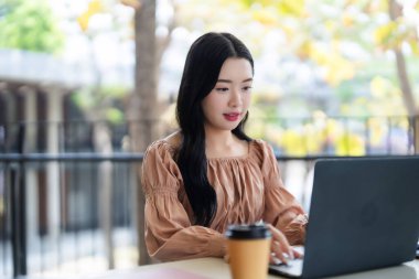 Genç Asyalı yetişkin kadın dışarıda taşınabilir bilgisayarına konsantre oluyor. Uzaktan kumandayla bir fincan kahveyle çalışıyor. Bu, modern esnek iş yerlerini gösteriyor.