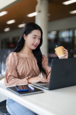 Gülümseyen Asyalı yetişkin kadın laptopta çalışıyor kafede kahvenin tadını çıkarıyor. Modern uzaktan çalışma dijital öğrenme ve üretkenlik vurgulanmıştır.