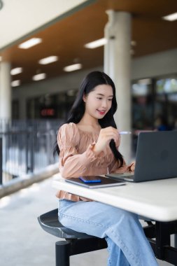 Gülümseyen Asyalı kadın modern bir binanın dışında dizüstü bilgisayarla ilgileniyor. Eğitim veya iş birliği için verimli faaliyet.