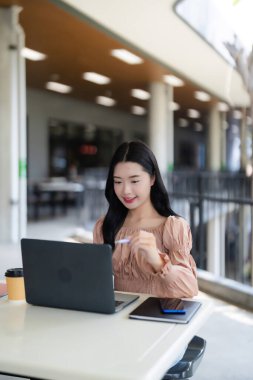 Gülümseyen Asyalı yetişkin kadın dışarıda dizüstü bilgisayarda çalışıyor. Dijital görevleri, öğrenmeyi, içerik yaratmayı ya da parlak bir kafeden iş yürütmeyi seviyor..