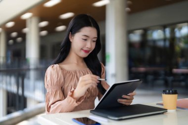 Yetişkin Asyalı kadın modern kafede dijital tablet kalem kullanıyor. Konsantre öğrenci profesyonel çalışmaları çevrimiçi içerik yaratıyor. Parlak davet çalışma alanı üretkenlik yeniliğine ilham verir.