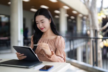 Asyalı yetişkin kadın aktif olarak dijital tablet stilus kalem açık hava kafesi kullanıyor. Verimli uzaktan çalışma ya da yaratıcı çalışma için modern ortam.