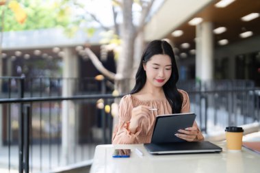 Genç Asyalı kadın stilus kalem kullanarak dijital tabletine konsantre oluyor. Çalışıyor ya da uzaktan çalışıyor. Modern bağlantıların tadını çıkarıyor..
