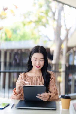 Uzaktan kumandalı kafede dijital tablet ve stil kullanan odaklanmış genç Asyalı kadın teknoloji üretkenliğinin keyfini çıkarıyor.