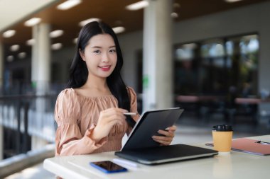 Genç Asyalı kadın, eğitim veya iş arayışında olan parlak çağdaş bir kafe ortamında dijital tablet ve stilus kullanıyor.