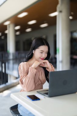 Modern eğitim ortamında veya çalışma alanında kulaklık takan mutlu Asyalı genç kadın dizüstü bilgisayarla meşgul gülümsüyor..