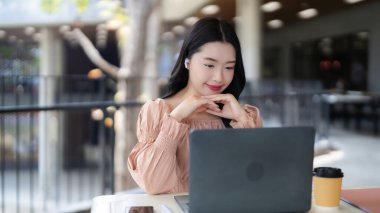 Odaklanmış Asyalı kadın canlı bir kafede dizüstü bilgisayarla çalışıyor. Genç profesyonel, kablosuz kulaklıklar ve kahve ile uzak verimliliği seviyor.