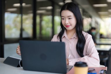 Genç Asyalı yetişkin kadın, kulaklıkları internet üzerinden etkinlik, uzaktan çalışma ya da çalışma oturumlarının keyfini çıkaran çağdaş kafe ortamında dizüstü bilgisayara odaklandı.