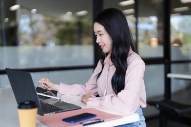 Genç Asyalı yetişkin kadın, modern, parlak bir çalışma alanında kulaklık takan bir stille dizüstü bilgisayar ve tablet kullanarak dijital çalışmaya odaklandı..