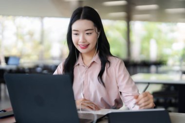 Genç Asyalı kadın diz üstü bilgisayar tableti ve kulaklıklarını kullanarak mutlu bir şekilde çalışıyor. Parlak bir çalışma alanındaki bir göreve odaklanıyor..