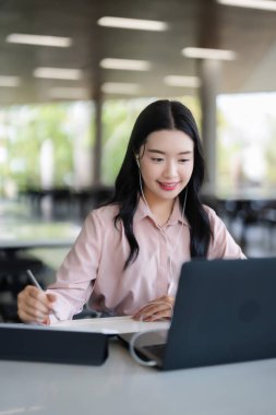Gülümseyen Asyalı yetişkin kadın dijital yaratıcıya odaklandı. Uzaktan çalışan, çevrimiçi modern kafe ortamını öğreniyor. Kulaklık takıyor, bilgisayarlı kalem kullanıyor.