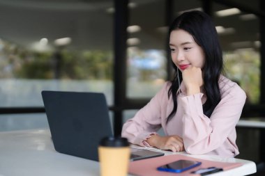 Genç Asyalı yetişkin, modern bir çalışma alanında dizüstü bilgisayar ekranı izlemekle meşgul. Gülümsüyor ve kulaklıklarla gizliliğin tadını çıkarıyor.