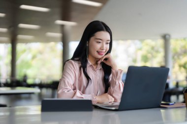 Gülümseyen genç Asyalı yetişkin kadın bilgisayarına odaklanmış kulaklık takıyor ve uzak işlerden ya da modern bir çevrede online eğitimden keyif alıyor.