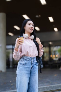 Yetişkin Asyalı kız öğrenci sabah kahvesini dışarıda içerek eğleniyor. Sırt çantasında kulaklık taşıyan günlük kıyafetler giyiyor. Parlak şehir arkaplanı.