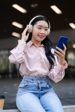 Gülümseyen Asyalı kadın kablosuz kulaklık takıyor dışarıda akıllı telefon içeriklerini seyrediyor. Modern teknolojiyle rahat anların tadını çıkarıyor..