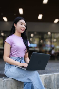 Genç Asyalı kadın dışarıda dizüstü bilgisayar yazarken gülümsüyor. Yetişkin öğrenci uzak çalışma dijital göçebe çevrimiçi eğitim odaklı