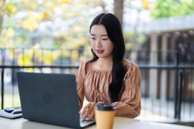 Yetişkin bir Asyalı kadın bilgisayarını bilgisayarla uzaktan çalışma ya da açık havada kişisel proje için kullanıyor. Üretken dijital yaşam tarzı.