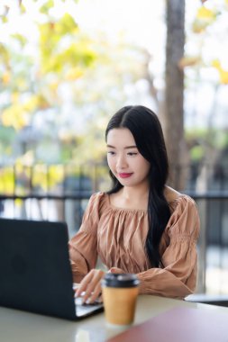 Yetişkin bir Asyalı kadın modern bir kafede dizüstü bilgisayar üzerinde çalışıyor. Açık hava doğal ışık arka plan bulanıklığı üretkenliğe odaklanmayı artırır. Etkili uzaktan çalışma.