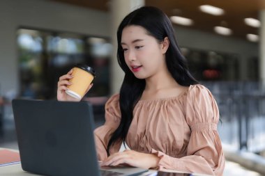 Genç Asyalı profesyonel bir kadın dizüstü bilgisayarda yazı yazıyor. Parlak, modern bir kafede kahvenin tadını çıkarıyor. Uzaktan çalışan online iş kadınlarını inceliyor.